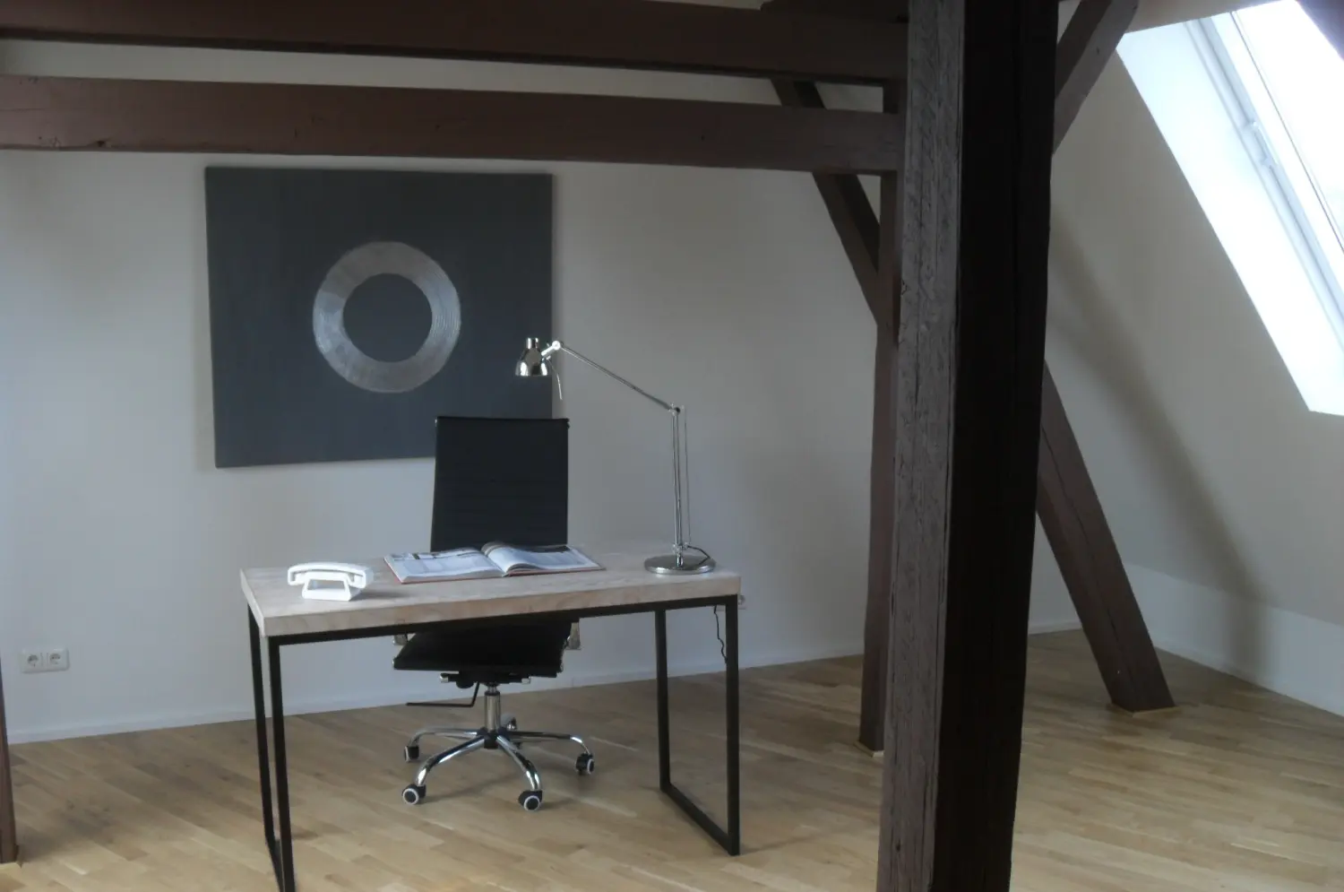 Fierle Baubetreuung – ein Arbeitsplatz in einem Dachzimmer mit Schreibtisch, Stuhl und einem Gemälde im Silberrahmen