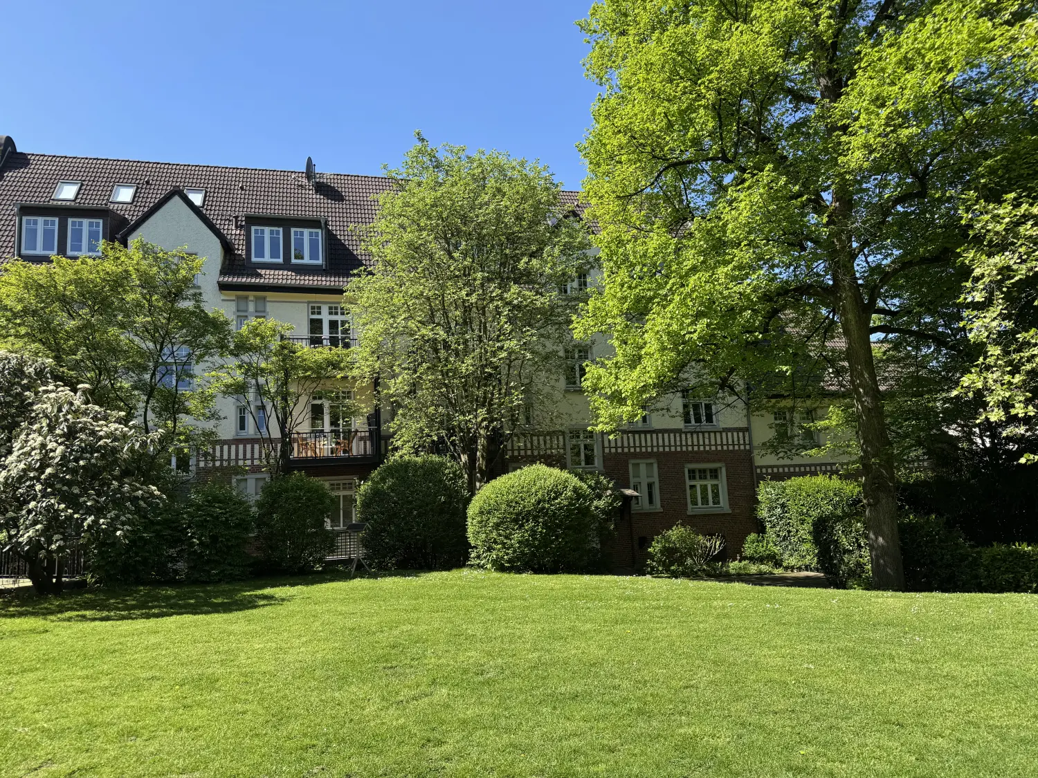 Fierle Baubetreuung – Wohnhaus mit heller Fassade und dunklem Dach, teilweise bedeckt von üppig grünen Bäumen und Sträuchern